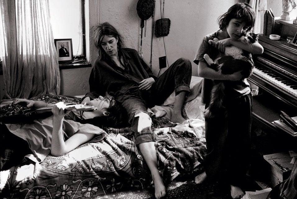 Singer Patti Smith sits on a bed as one son lies back, holding a guitar and another son stands, holding a cat.