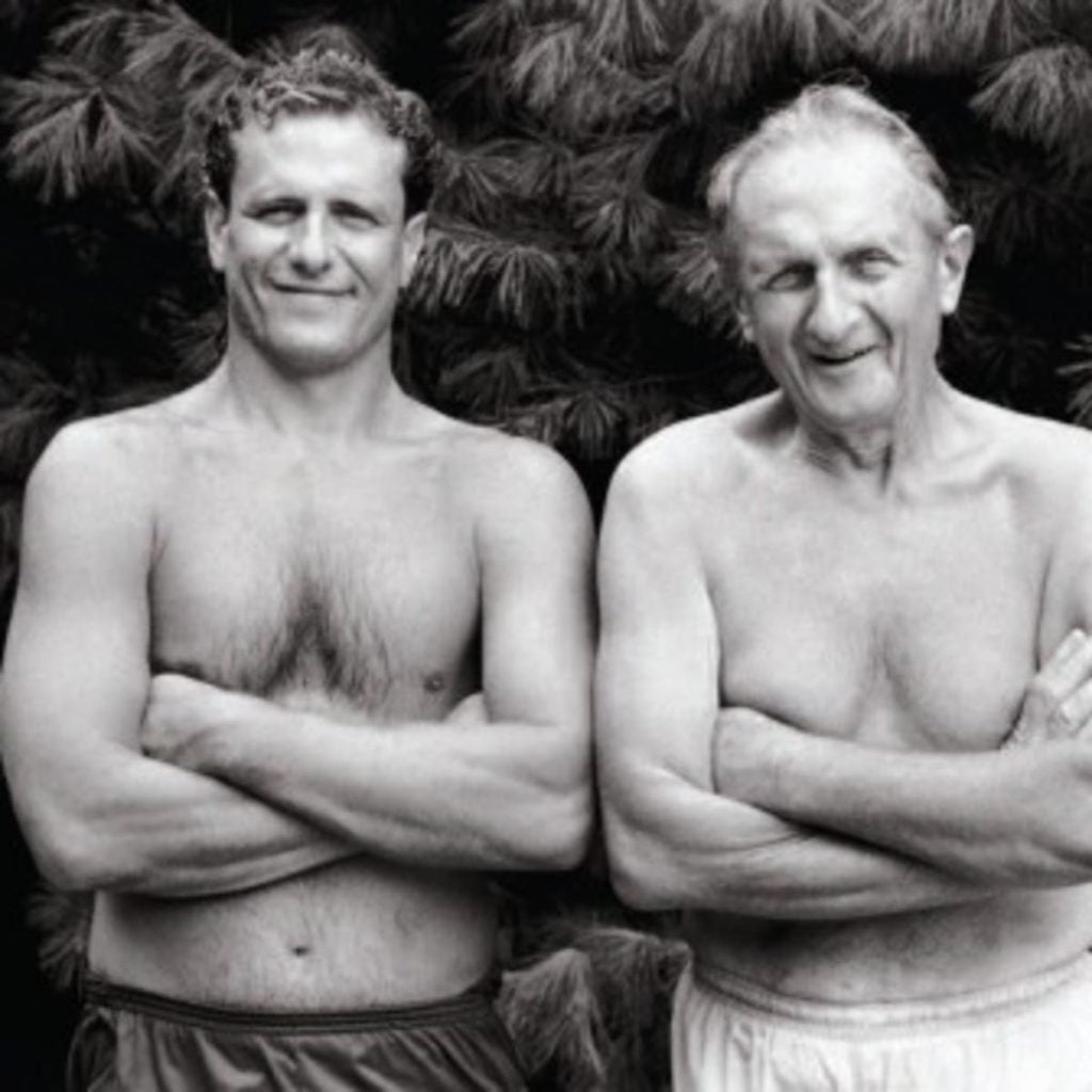Two shirtless men stand side by side, both with their arms crossed.