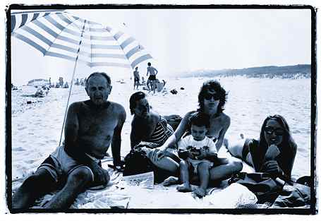 One man and three women sit in a semi-circle on a beach under a striped umbrella. The woman one spot from the right holds a baby.