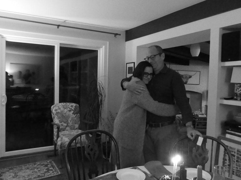 A man and woman hug, standing, behind a table with a birthday cake.