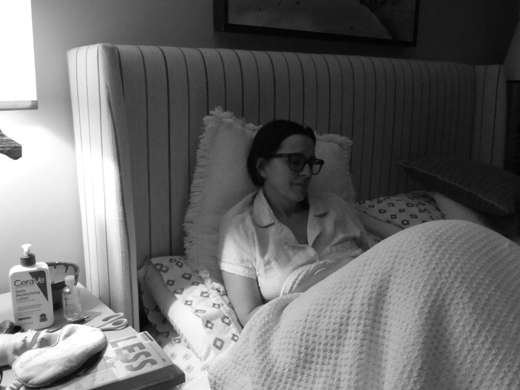 A woman sits upright in bed, eyes closed