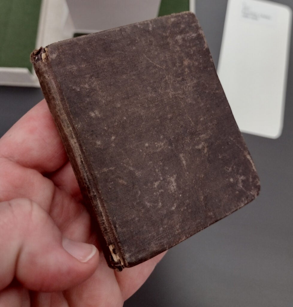 From a bird's eye view, there is a white human hand positioned close to the camera with the palm facing upward. There is a small, brown, three by two and a half inch booklet placed on top of the fingers on the hand. The book has no words or pictures on it, but appears to be old.